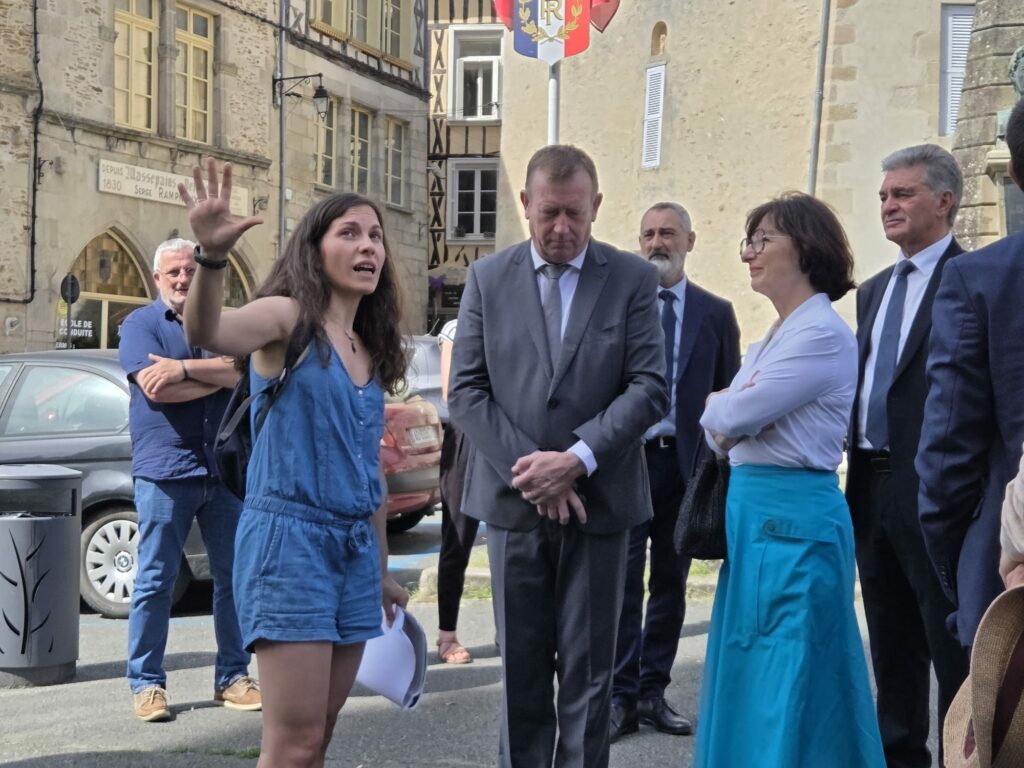 Guide du patrimoine présentant la Collégiale de Saint-Léonard de Noblat au Maire Alain Darbon, à la Préfète de Région Sophie Brocas et à leur délégation dans une rue du centre-ville.